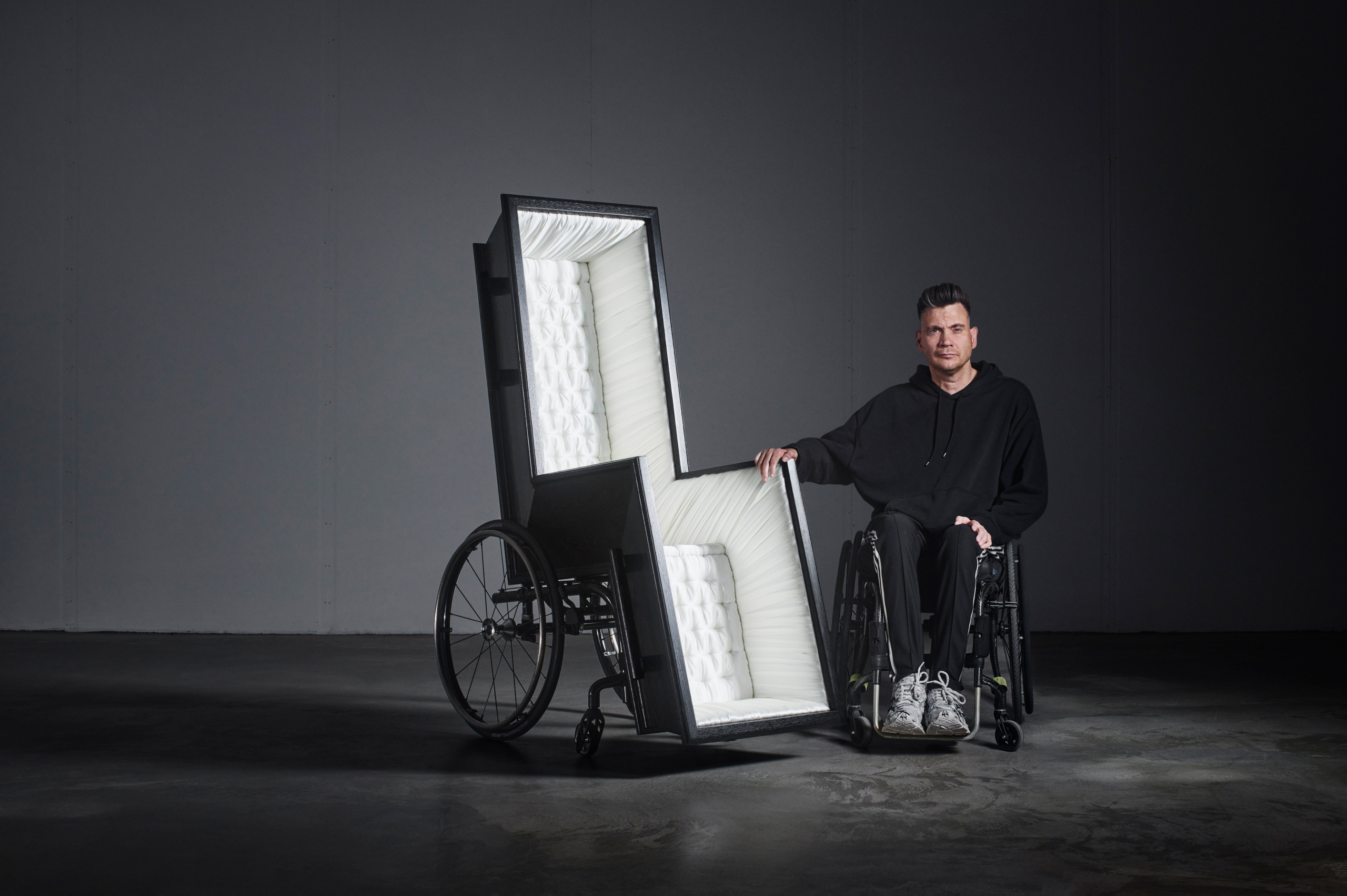 Man i rullstol håller i ett konstverk som är en mix av en rullstol och en likkista. Studiomiljö.