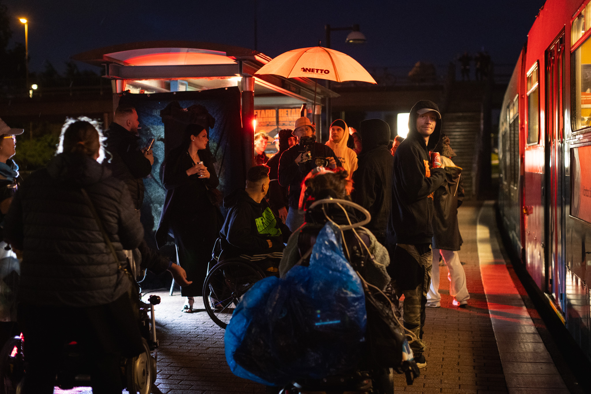 Natt, ljuset är färgat orange av lampor, många personer står på en spårvagnshållplats, feststämning, en spårvagn står på hållplatsen.