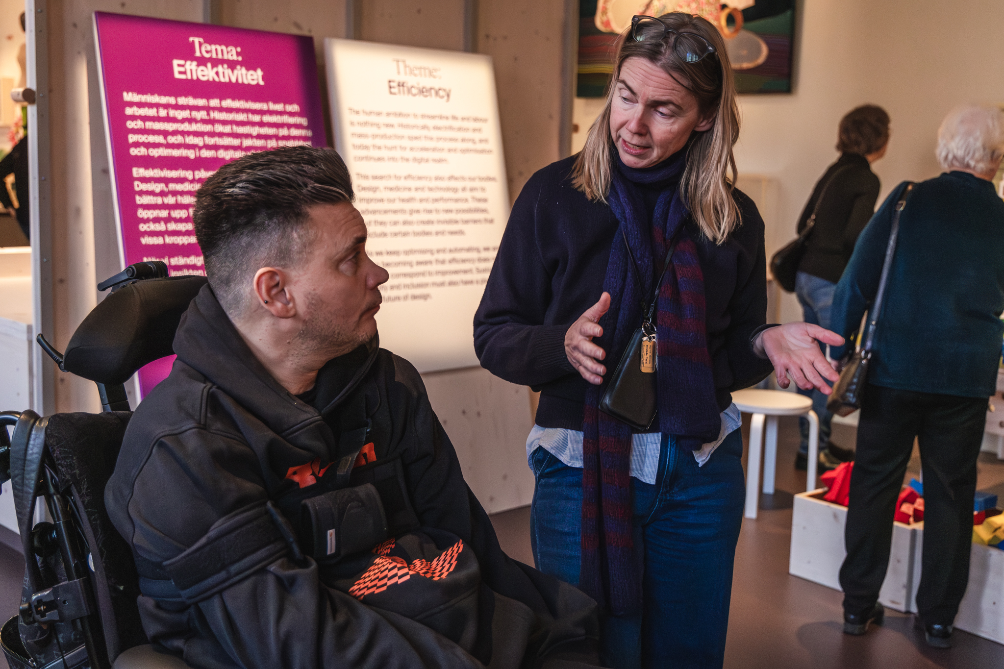 Jessica gestikulerar och Anders i rullstol tittar intresserat, från museimiljö